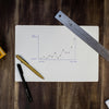 A hand-drawn line graph on paper with a metal ruler and two pens