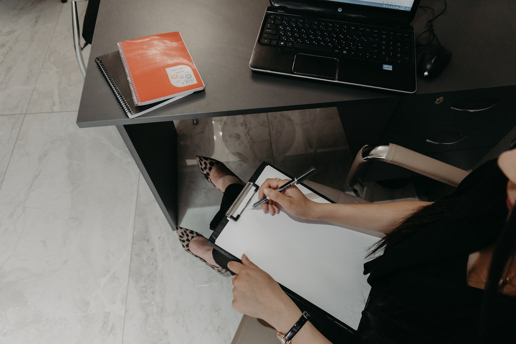 Person writing on clipboard at desk with laptop
